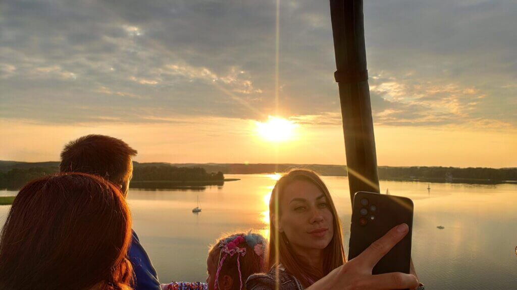 A group of people in a hot air balloon taking a picture of the sun setting over a lake.